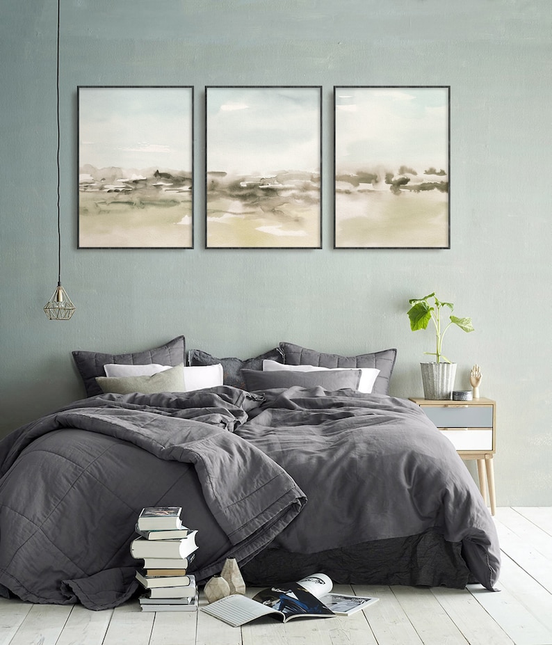 Op de afbeelding: Drie ingelijste abstracte aquarellen in gedempte groene en bruine tinten, hangend aan een lichtblauwe muur boven een grijs bed met een grijs dekbed en witte kussens.