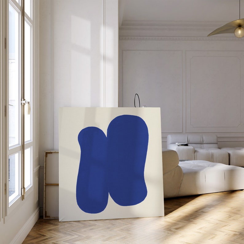May include: A white canvas with two abstract blue shapes, one larger than the other, on a white wall in a room with a white sofa.