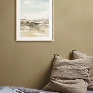 Op de afbeelding: Een ingelijst aquarel schilderij van een landschap met een lichtblauwe lucht, bruine en grijze heuvels en een lichtgele voorgrond. Het schilderij hangt aan een lichtbruine muur boven een bed met twee beige kussens en een gele mok op een houten dienblad.