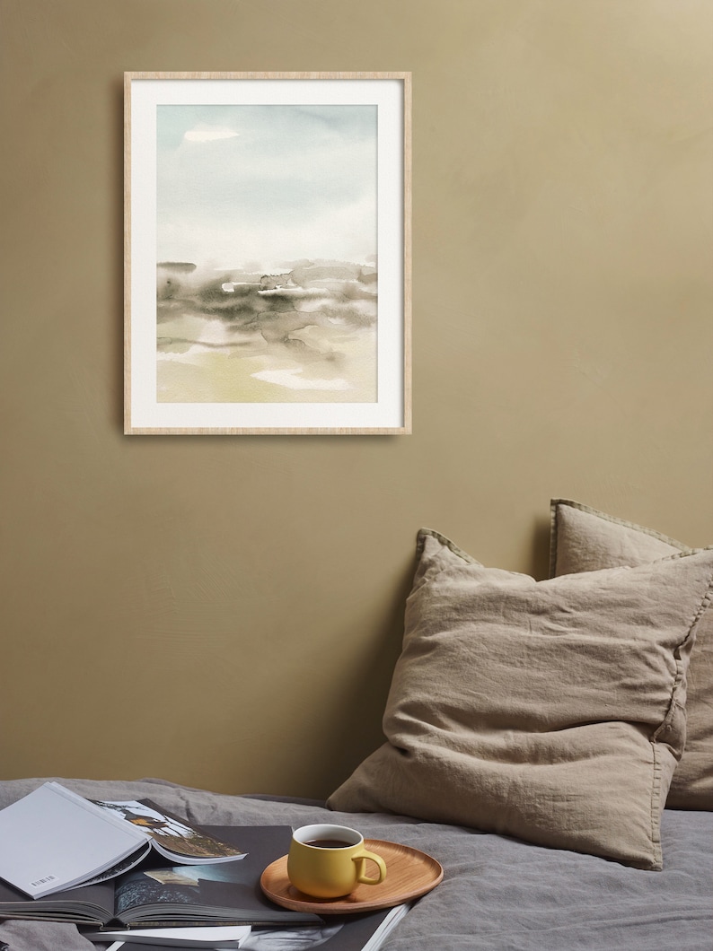 Op de afbeelding: Een ingelijst aquarel schilderij van een landschap met een lichtblauwe lucht, bruine en grijze heuvels en een lichtgele voorgrond. Het schilderij hangt aan een lichtbruine muur boven een bed met twee beige kussens en een gele mok op een houten dienblad.