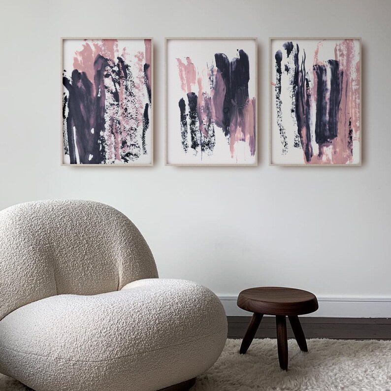May include: Three abstract paintings in white frames with pink and purple brushstrokes on a white background. The paintings are hanging on a white wall above a white fluffy armchair and a wooden stool.