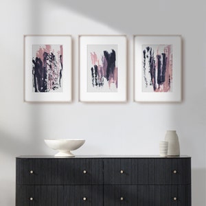 May include: Three framed abstract paintings with pink and purple brushstrokes on a white background. The paintings are hanging on a white wall above a black dresser with gold handles.