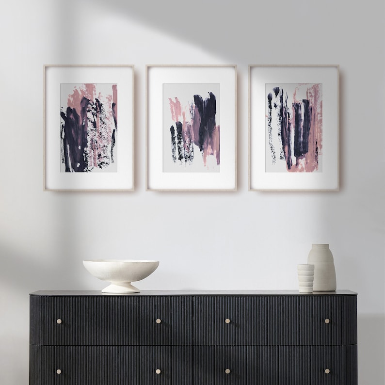 May include: Three framed abstract paintings with pink and purple brushstrokes on a white background. The paintings are hanging on a white wall above a black dresser with gold handles.