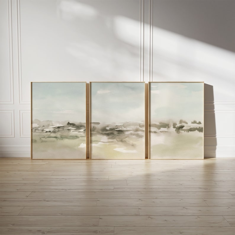 Op de afbeelding: Een set van drie abstracte landschapschilderijen in gedempte tinten groen, blauw en bruin. De schilderijen zijn ingelijst in goud en hangen aan een witte muur.