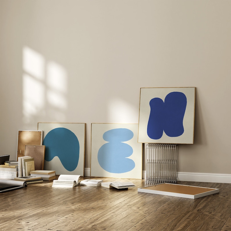 May include: Three framed abstract art prints with blue and light blue shapes on a white background. The prints are hanging on a light beige wall.