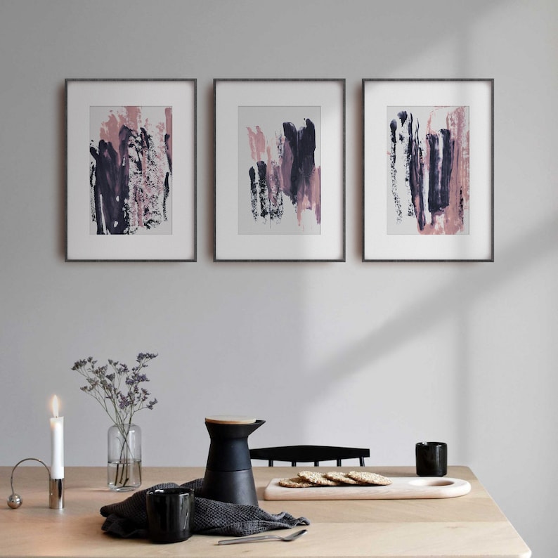 May include: Three framed abstract art prints with pink and purple brushstrokes on a white background. The frames are black and the prints are hung on a white wall.