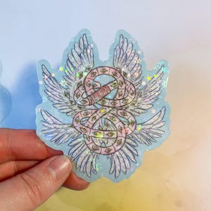 May include: A holographic sticker featuring a stylized angel with multiple eyes and wings. The design includes a pink and white serpent-like form, surrounded by white wings and small stars, all set against a light blue background.
