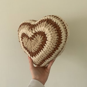 Könnte beinhalten: Ein gehäkeltes Herzförmiges Kissen in Braun und Creme. Das Kissen ist aus weichem, flauschigem Garn gefertigt und hat ein strukturiertes, gewebtes Design.