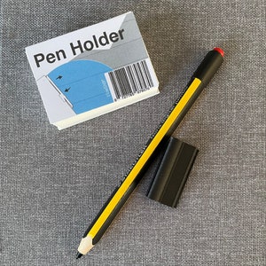 Könnte beinhalten: Ein schwarz-gelber Bleistift mit rotem Radiergummi, ein schwarzer Radiergummi und ein weiß-blauer Stifthalter mit dem Text "Pen Holder" liegen auf einer grauen Oberfläche. Der Stifthalter hat einen Barcode.