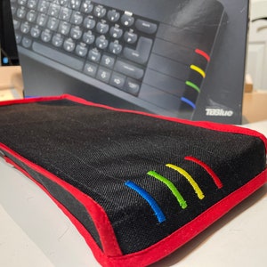 Peut inclure: Un étui en tissu noir avec une bordure rouge et cinq lignes brodées colorées. L'étui est conçu pour contenir un clavier.
