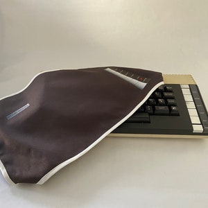 Puede incluir: Teclado de ordenador Atari 800XL con una funda marrón. La funda tiene un ribete blanco y el texto "ATARI 800XL". El teclado tiene teclas negras y una parte superior beige con teclas de función blancas.