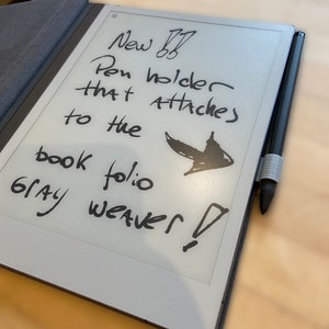 Puede incluir: Primer plano de un lector electrónico gris con una nota escrita a mano que dice "Nuevo portaplumas que se adhiere al folio de libro gris tejedor!" La nota también incluye un pequeño dibujo de una flecha.