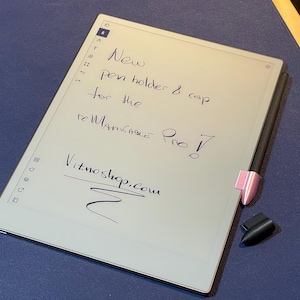 Könnte beinhalten: Ein weißer E-Reader mit einem schwarzen Stift und einem rosa Stifthalter. Der Bildschirm zeigt handschriftlichen Text, der "New pen holder & cap for the Remarkable Pro! Vitmoshop.com" lautet.