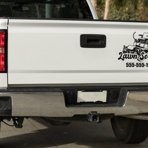 May include: White pickup truck with a black and white lawn service logo on the tailgate. The logo features a lawnmower and the text "Lawn Service" with the phone number 555-555-1212.