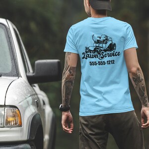 May include: A light blue t-shirt featuring a black lawnmower graphic and the text "Lawn Service" with a phone number. The person is wearing the shirt and standing in front of a white truck.