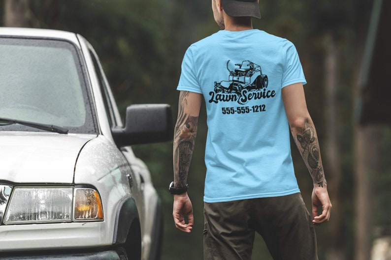 May include: A light blue t-shirt featuring a black lawnmower graphic and the text "Lawn Service" with a phone number. The person is wearing the shirt and standing in front of a white truck.