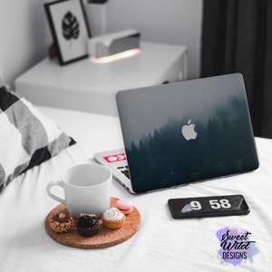 May include: A white ceramic mug with a white laptop computer with a forest scene on the screen. The laptop is on a white bed with a black and white checkered pillow. There are small pastries on a cork coaster next to the mug. A black cell phone with a digital clock displaying 9:58 is on the bed.