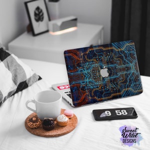 Puede incluir: Un ordenador portátil con un diseño de placa de circuito azul, naranja y amarillo en la tapa. El portátil está abierto y sentado en una colcha blanca con una taza blanca y un plato de pasteles. La hora es 9:58 en un teléfono negro.