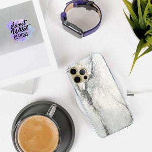 May include: A white marble phone case with a gold camera lens cover. The case is on a wireless charging pad. The case is on a white surface with a cup of coffee and a watch.
