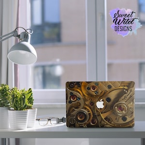 Könnte beinhalten: Ein Laptop mit einem goldenen und braunen Zahnrad-Design auf der Abdeckung. Das Design zeigt eine Nahaufnahme eines Uhrwerks mit komplizierten Details. Das Apple-Logo ist auf der Abdeckung sichtbar.
