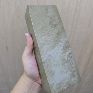 Può includere: Un blocco di pietra rettangolare grigio chiaro con una superficie strutturata. Il blocco ha una forma trapezoidale ed è tenuto in mano. La pietra ha un aspetto invecchiato.
