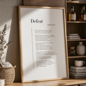 Puede incluir: Una impresión enmarcada con la palabra "Defeat" y un poema de Kahlil Gibran. La impresión tiene un marco de madera clara y se exhibe en un mueble de madera. También se ven una cesta de mimbre, dos tazas blancas y una planta decorativa.
