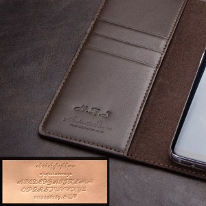 Puede incluir: Una funda de teléfono de cuero marrón con ranuras para tarjetas, con el texto en relieve "N.K.S Artisan Atelier Handmade leather works". La funda está abierta, revelando un smartphone. Una pequeña inserción muestra muestras de alfabeto y números.