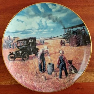 Puede incluir: Plato decorativo con una escena rural que muestra un coche antiguo, un tractor y figuras en un campo. El plato tiene un borde dorado y representa una escena de cosecha con un perro y dos personas cerca de un barril. El cielo es azul con nubes.