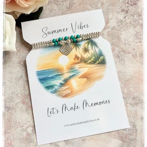 May include: A silver bracelet with turquoise beads and a diamond-shaped charm is presented on a card. The card reads "Summer Vibes" and "Let's Make Memories", and features a beach scene with palm trees.