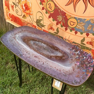 May include: A purple and brown resin table with a unique, swirling design. The table has a gold rim and sits on black hairpin legs.