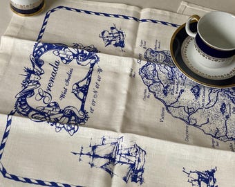 May include: A cream linen table runner featuring a blue map of Grenada, West Indies, and sailing ships. The runner has a blue rope border and text that reads "Grenada, West Indies". A teacup and saucer with blue and gold trim are on the runner.