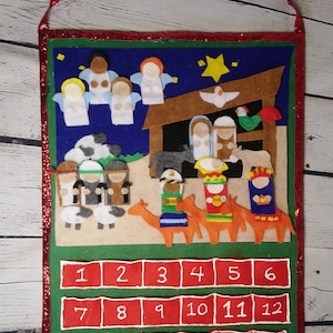 Peut inclure: Un calendrier de l'avent de Noël en feutre avec une scène de la nativité et une étoile au sommet. Les chiffres de 1 à 24 sont affichés en blanc sur des carrés rouges.