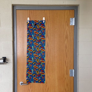 May include: A blue fabric door banner with a repeating pattern of red and yellow "Super" and "Wow" words with white outlines and yellow starbursts. The banner is hanging on a wooden door with a silver door handle.