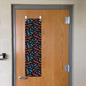 May include: A black door with a colorful feather pattern door hanger. The hanger is attached to the door with two white clips.
