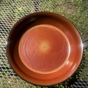 May include: A round ceramic bowl with a smooth, glossy interior featuring a gradient of deep burnt orange and reddish-brown tones. The center of the bowl is a lighter, warm golden-tan hue, transitioning into darker, rich earth tones toward the rim.