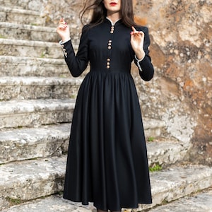 Peut inclure: Une robe noire à manches longues avec des boutons dorés et un col montant. La robe a un corsage ajusté et une jupe évasée. Les manches ont une bordure en dentelle blanche aux poignets. La personne porte des chaussures et des bas noirs. Un bandeau de perles est porté.