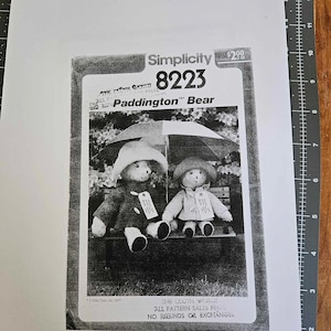May include: Black and white photo of a Simplicity sewing pattern for a Paddington Bear. The pattern number is 8223. The photo shows two Paddington Bear stuffed toys wearing raincoats and holding umbrellas.