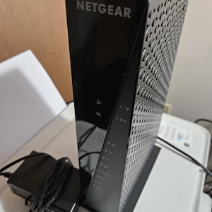 May include: A black NETGEAR router with a triangular design and a perforated side panel. The router has indicator lights and the NETGEAR logo in white. An AC adapter with a power cord is connected to the router.