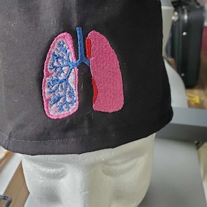 May include: Black scrub hat with an embroidered image of a pink and blue lung.