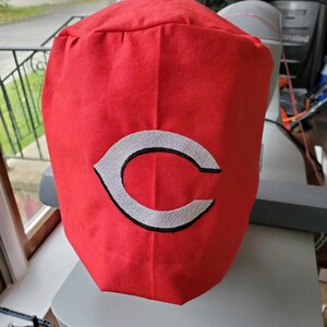 May include: A red fabric hat featuring a white and black embroidered "C" logo. The hat is made of a textured material and is likely a sports-themed accessory.