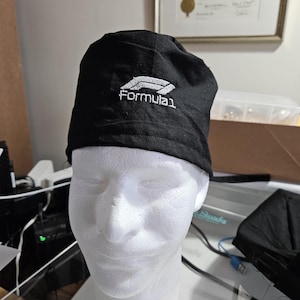 May include: A black scrub cap featuring the white embroidered Formula 1 logo on the front center. The cap is constructed from a solid black fabric and is displayed on a white mannequin head, showcasing the detailed stitching of the logo.