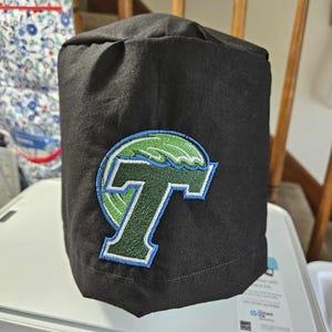 May include: A black scrub cap featuring an embroidered patch on the front. The patch displays a large dark green letter T with a light blue outline, positioned in front of a stylised light green wave graphic that curves around the top of the letter.