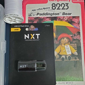 May include: A Simplicity sewing pattern for a Paddington Bear, featuring a colorful umbrella and a yellow raincoat. The pattern number is 8223. The pattern is for sale at The Cloth World, with a final sale price of $200.00.