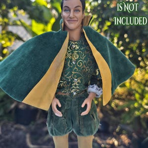 May include: A doll wearing a green and gold costume with a cape. The costume includes a green velvet cape with gold trim, a green and gold patterned top, and green velvet shorts. The text "DOLL IS NOT INCLUDED" is visible.