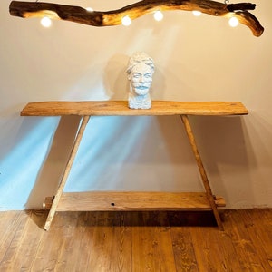 May include: A rustic wooden branch chandelier with four light bulbs hanging from the ceiling. The chandelier is lit and casts a warm glow. The chandelier is hanging above a wooden table with a white sculpture on top.