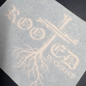 K&ouml;nnte beinhalten: Wei&szlig;er Vinyl-Aufkleber mit dem Wort "Rooted" in einer stilisierten Schriftart, darunter ein Kreuz und Baumwurzeln. Der Text "in Christ" befindet sich unter den Wurzeln.