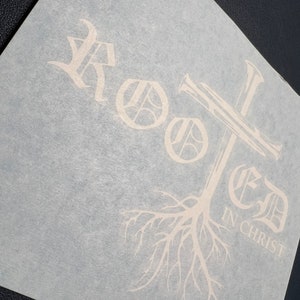 K&ouml;nnte beinhalten: Wei&szlig;er Vinyl-Aufkleber mit dem Wort "Rooted" in einer stilisierten Schriftart, einem Kreuz und einem Baumwurzel-Design. Der Text "In Christ" befindet sich unter den Baumwurzeln.
