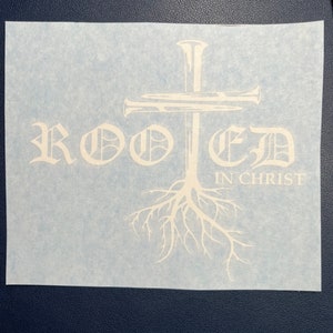 K&ouml;nnte beinhalten: Wei&szlig;er Vinyl-Aufkleber mit einem Kreuz aus N&auml;geln und dem Wort "ROOTED" in einer stilisierten Schriftart mit den Worten "IN CHRIST" darunter. Das Kreuz hat Wurzeln, die aus seiner Basis wachsen.