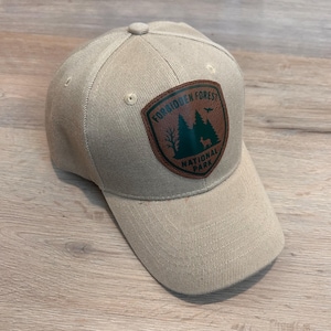 May include: Tan baseball cap with a curved brim and a button on top. The cap features a patch with the words "Forbidden Forest National Park" and a forest graphic. The cap is made of a durable fabric.