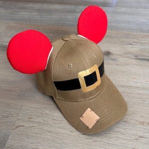 May include: A brown baseball cap with a black belt and gold buckle. The cap has two red fabric ears attached to the top.